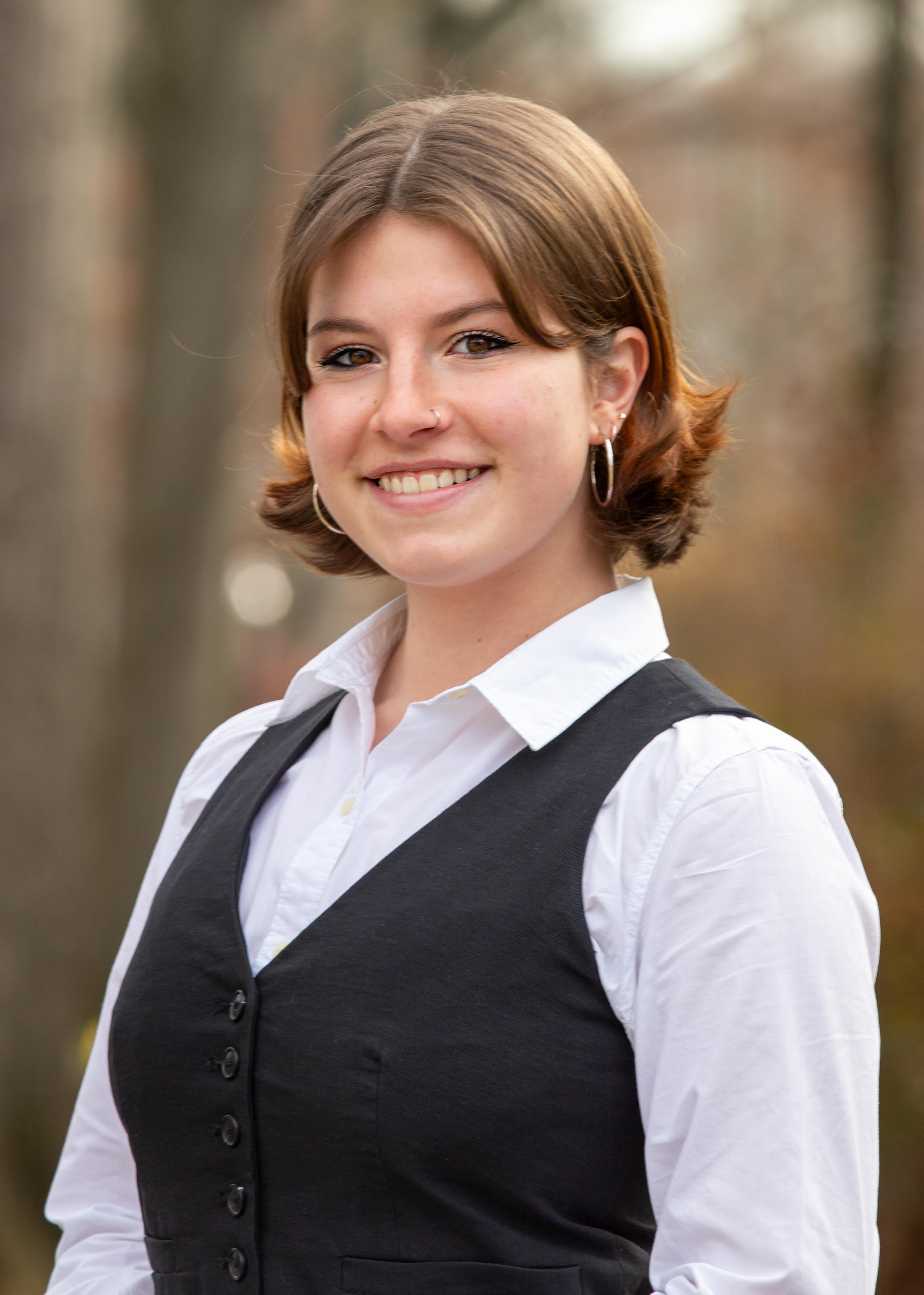 Anna has brown shoulder length hair and is wearing a white blouse and black vest, along with silver hoop earrings.