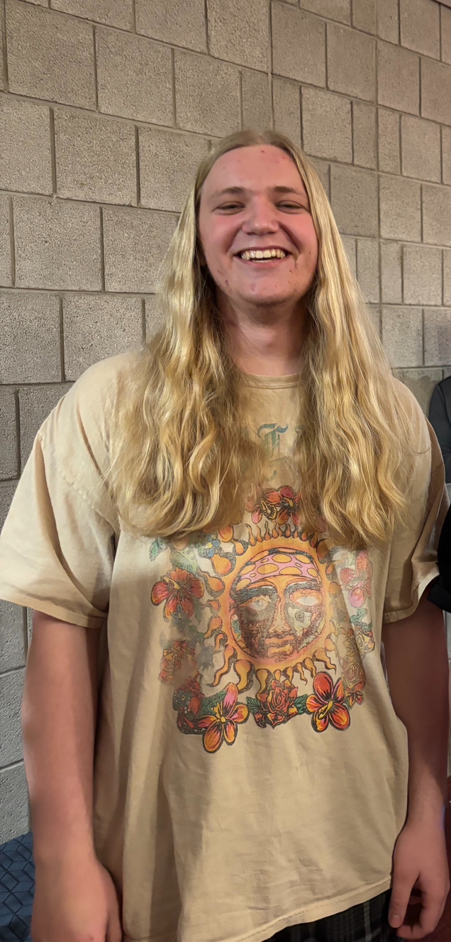 Smiling Max has long blond hair and is wearing a tan t-shirt while standing in front of a tan brick wall.