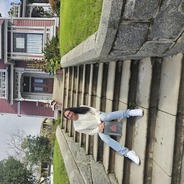 Ariel sitting in front of the red charmed house with light wash jeans and a beige long sleeve shirt with dark shiny straightened hair
