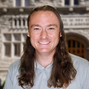 Scottie with long dark brown hair in a gray polo smiling in front of a gray building