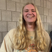 Smiling Max has long blond hair and is wearing a tan t-shirt while standing in front of a tan brick wall.