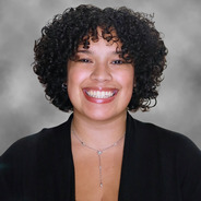 Mia in a black blouse in front of a gray and white background with medium length curly dark hair