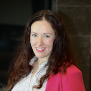 Natalie is pictured here. She is a fair-skinned woman with brown hair. She is wearing a white blouse with a pink blazer and pink lipstick.